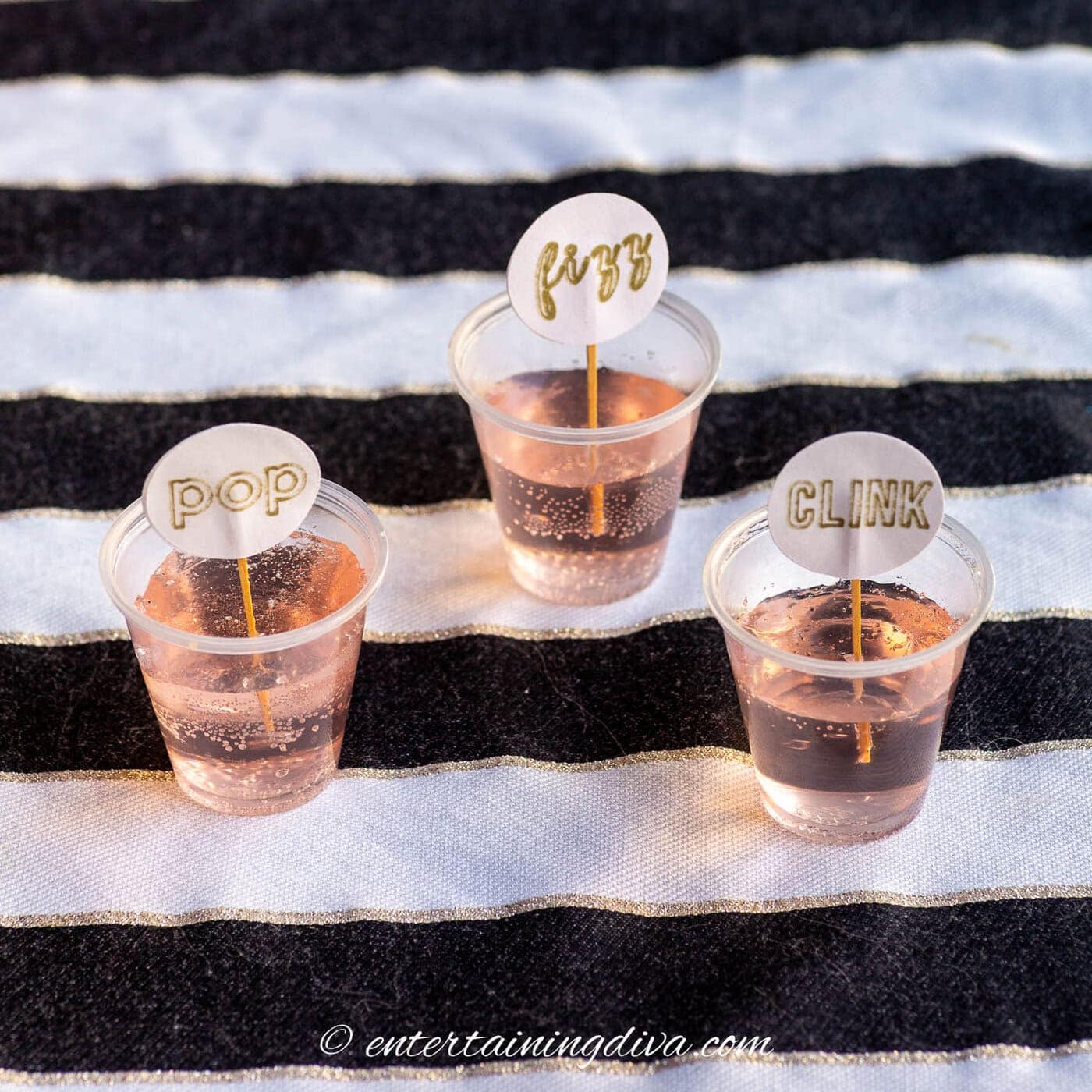 Three plastic cups with fizzy pink drinks sit on a striped cloth, each with a drink stirrer labeled "POP," "FIZZ," or "CLINK.