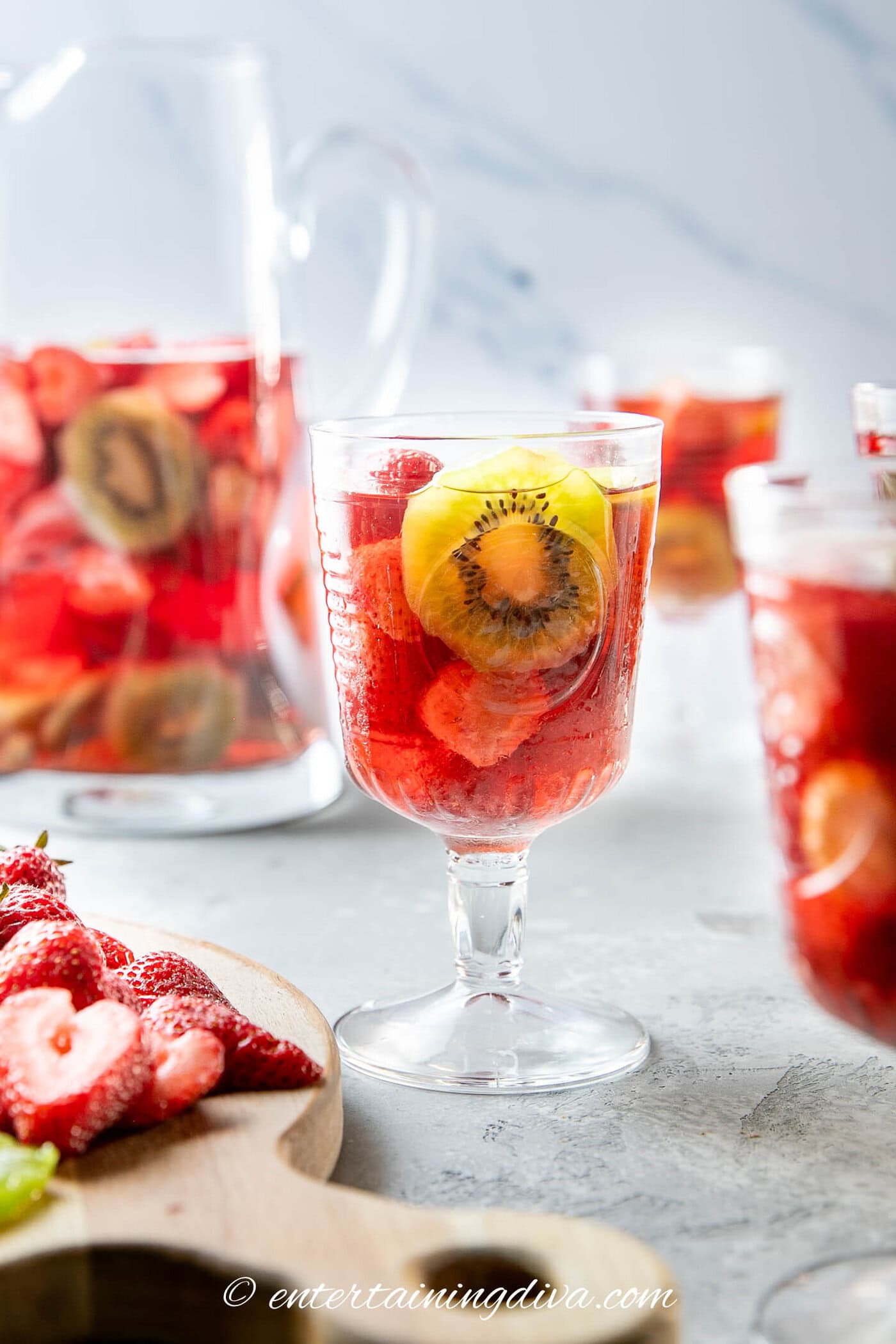 A glass of red fruit punch with sliced strawberries and kiwi, with a pitcher and more glasses in the background. Fresh fruit slices are on a wooden board in the foreground.