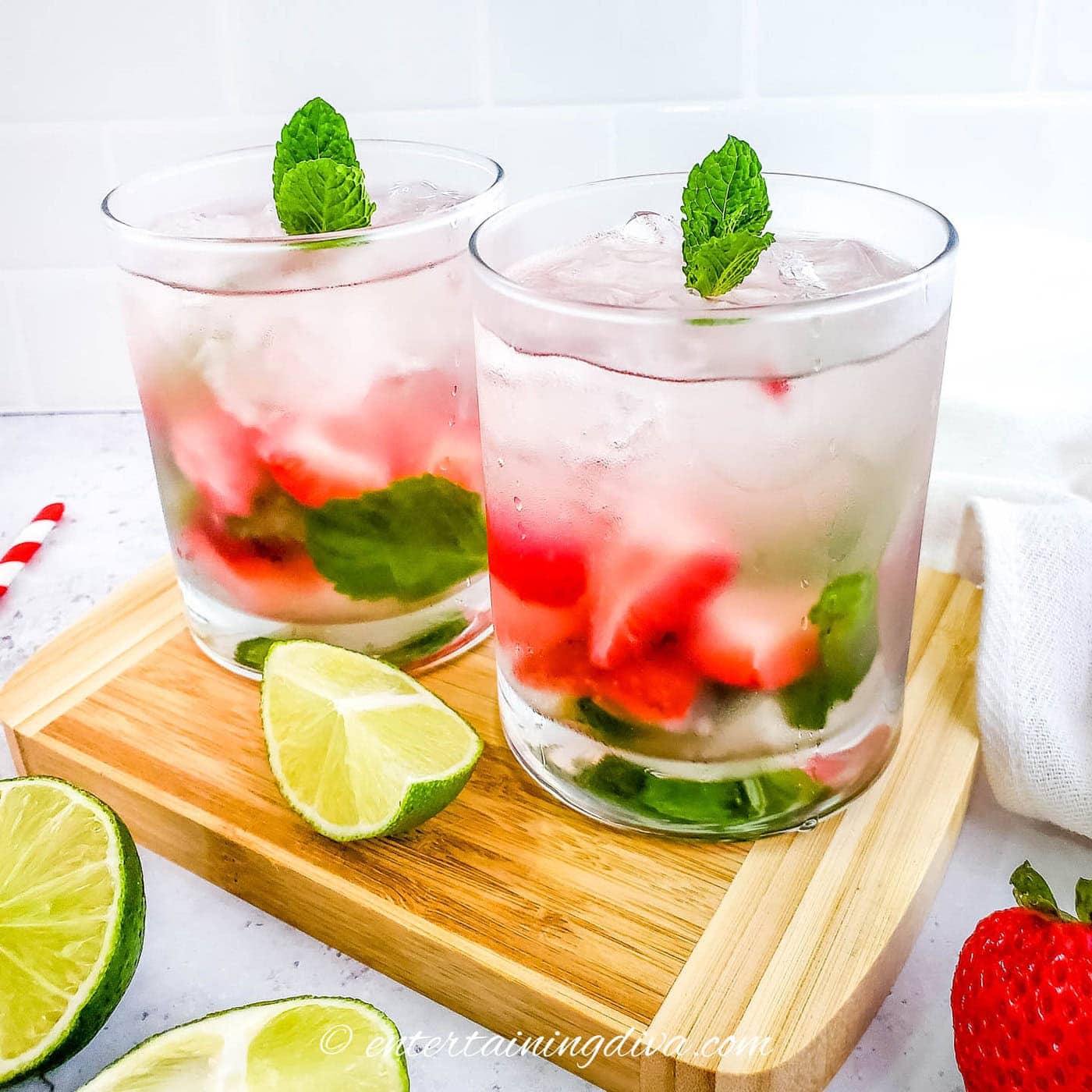 Two glasses of iced strawberry mojitos garnished with mint leaves, placed on a wooden tray with lime wedges and strawberries nearby.