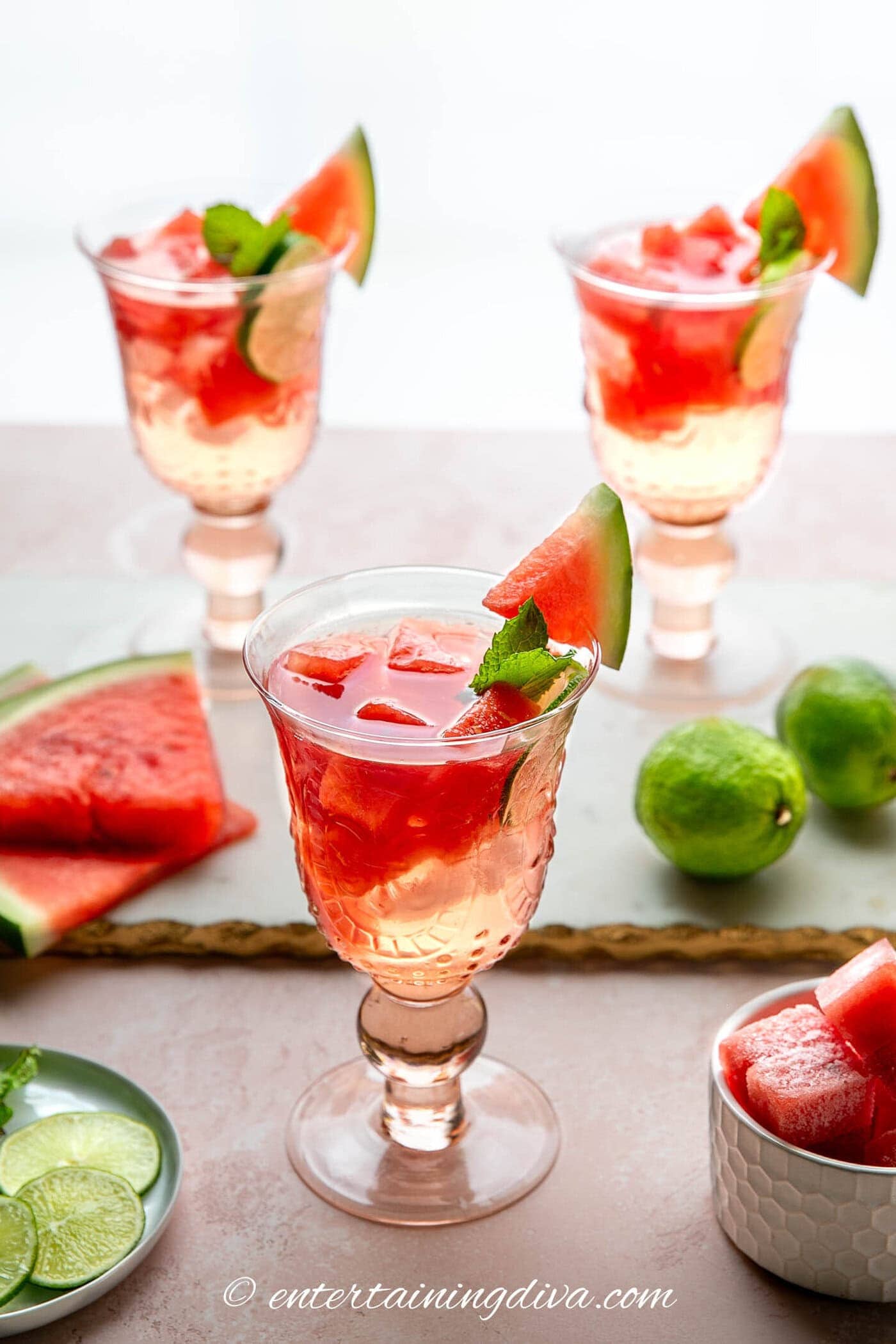 Three glasses of a pink drink garnished with watermelon cubes, lime slices, and mint on a table, with extra watermelon and limes in the background.
