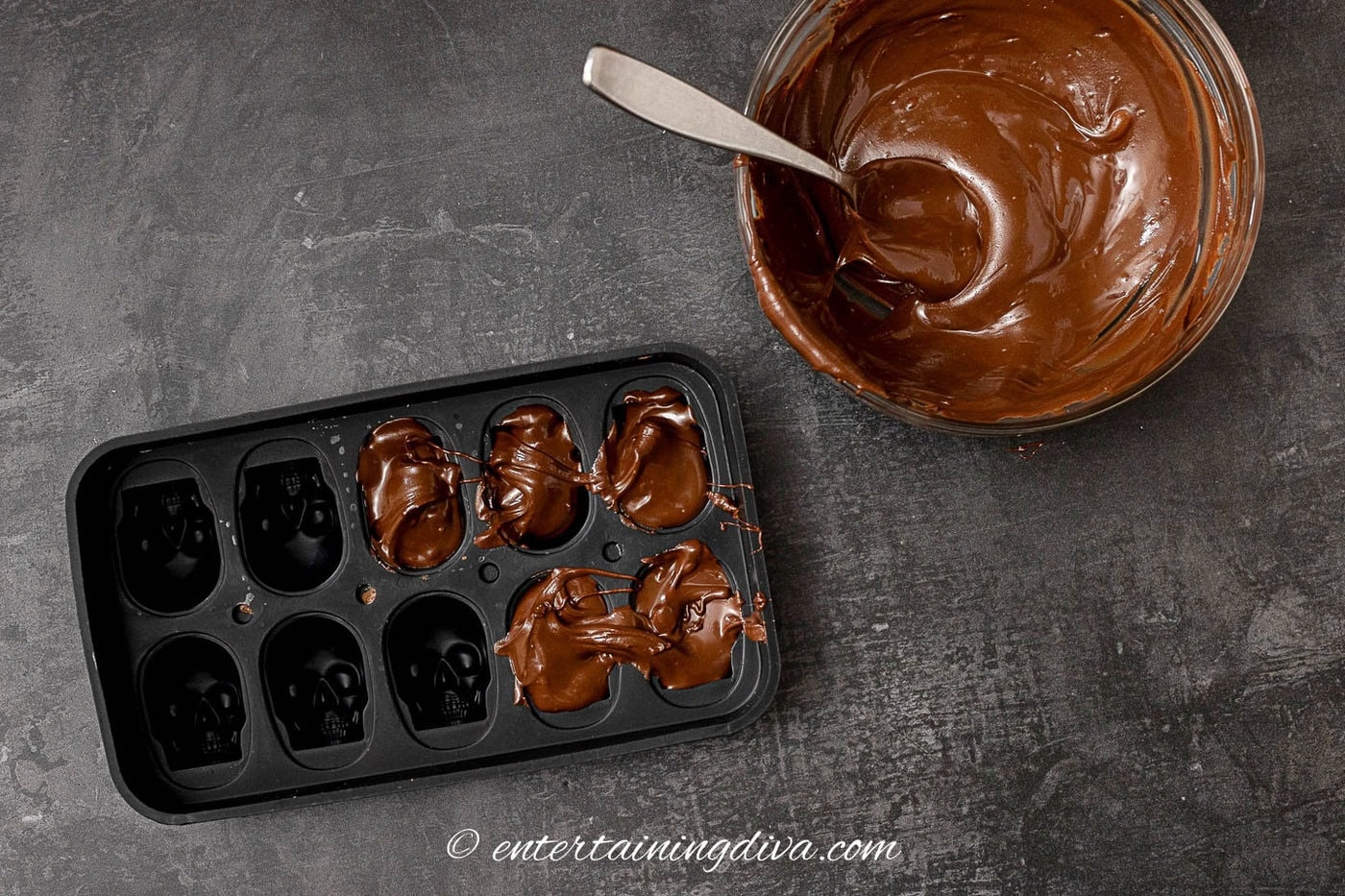 Melted chocolate chips in a bowl beside a skull mold that is half full of melted chocolate