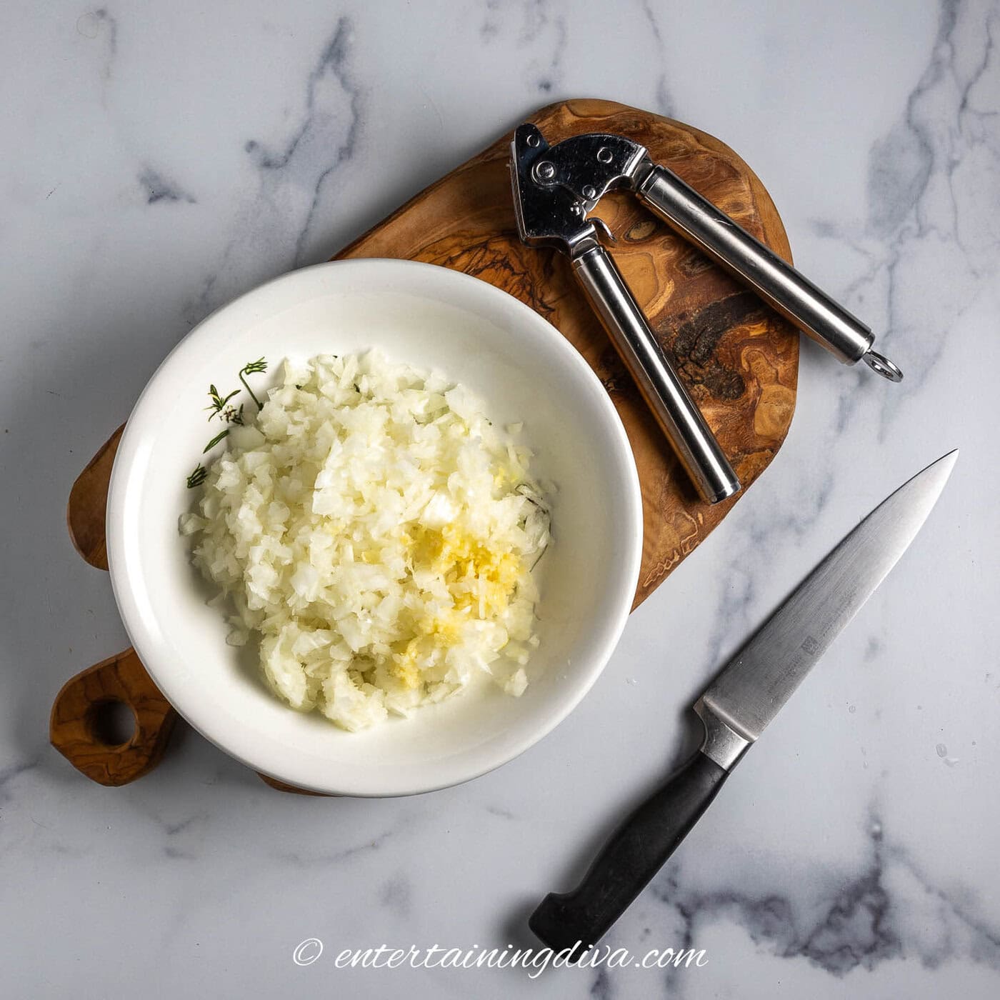 chopped onions and minced garlic in a bowl beside a garlic press and chefs knife