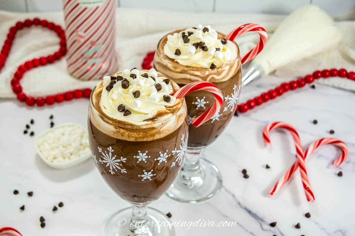 Two glasses of spiked hot cocoa in front of a peppermint vodka bottle with candy canes, and mini marshmallows around them