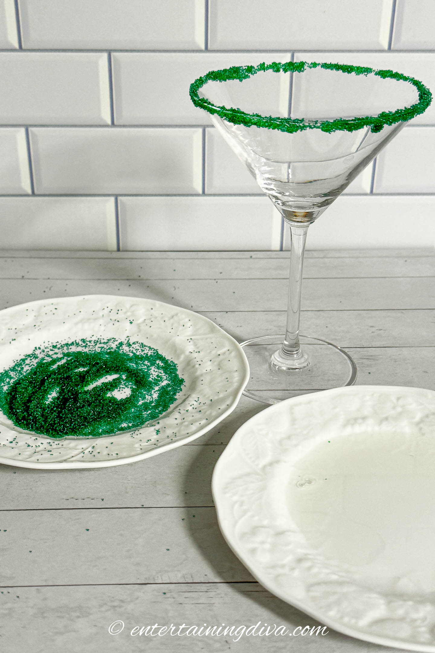 martini glass rimmed with green sugar sprinkles beside a plate covered with green sprinkles and one covered with simple syrup