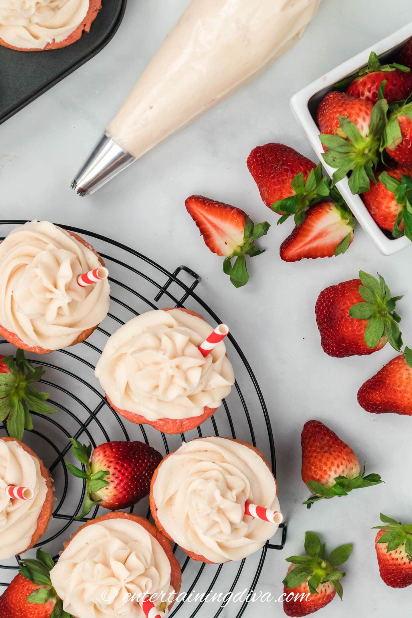 a piping bag full fo strawberry cream cheese frosting beside some strawberries and cupcakes