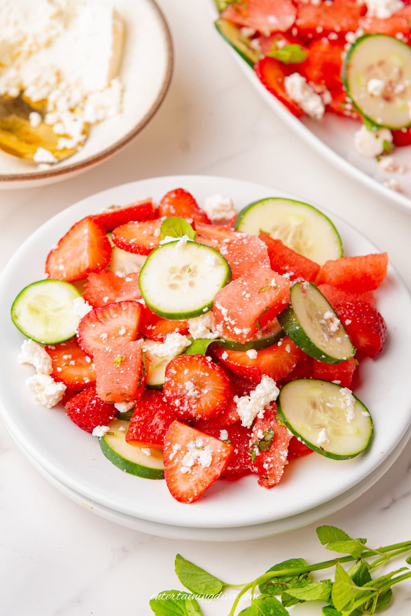 A plate of salad with sliced cucumbers, strawberries, watermelon, and crumbled feta cheese on a white surface.