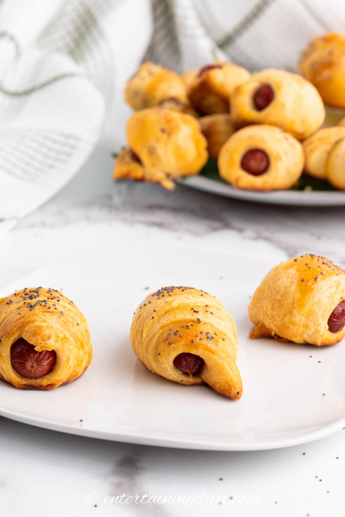 Three pigs in a blanket with poppy seeds on a white plate, with more pastries on a platter in the background.