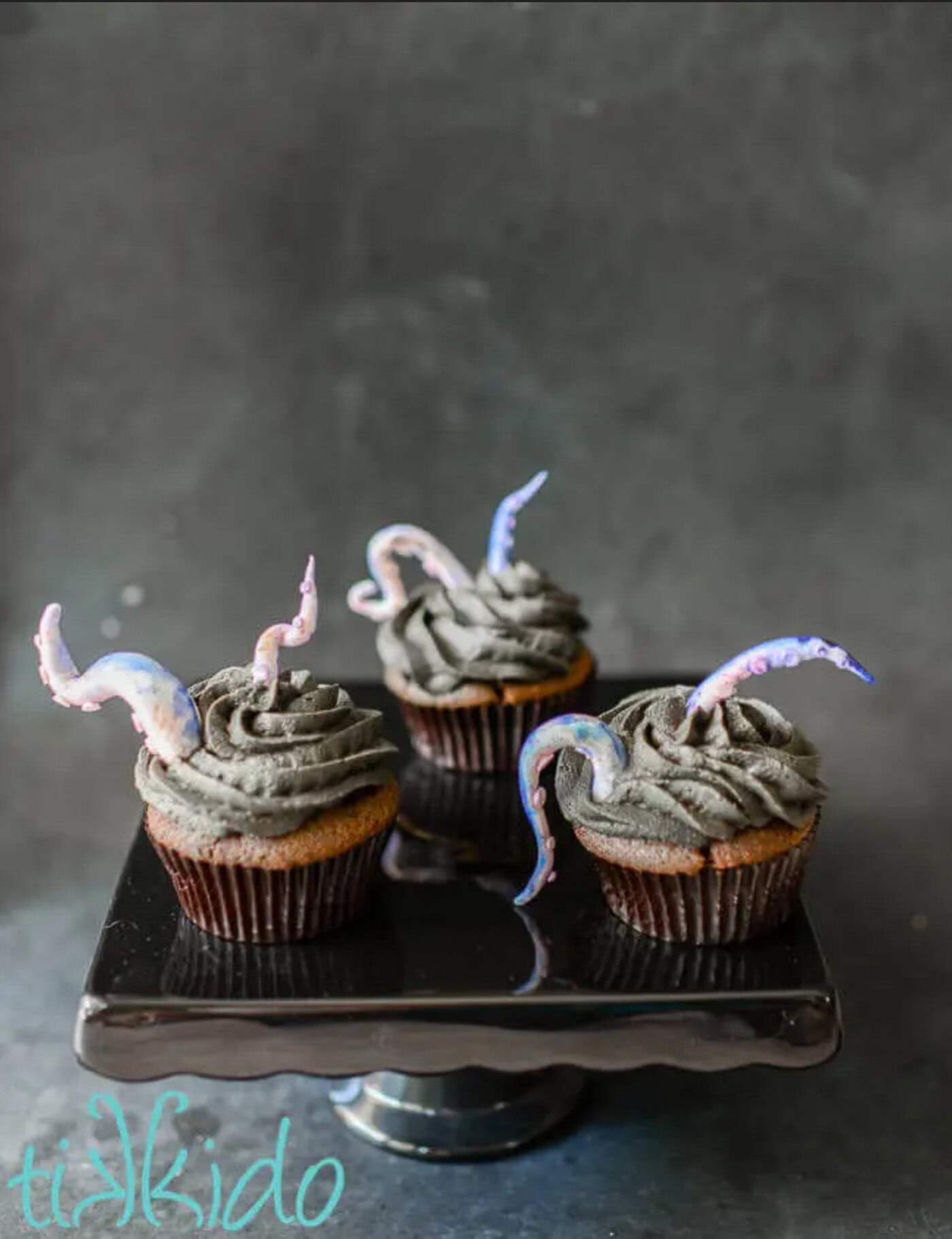 Three cupcakes with dark frosting are topped with decorative purple and pink tentacle shapes, displayed on a black cake stand against a dark background.