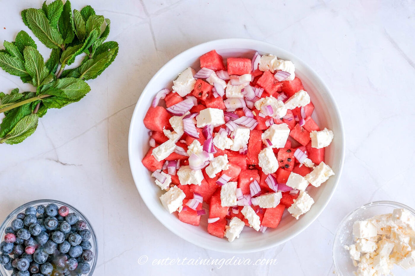 watermelon, feta and onions in a bowl with mint, blueberries and feta around the outside