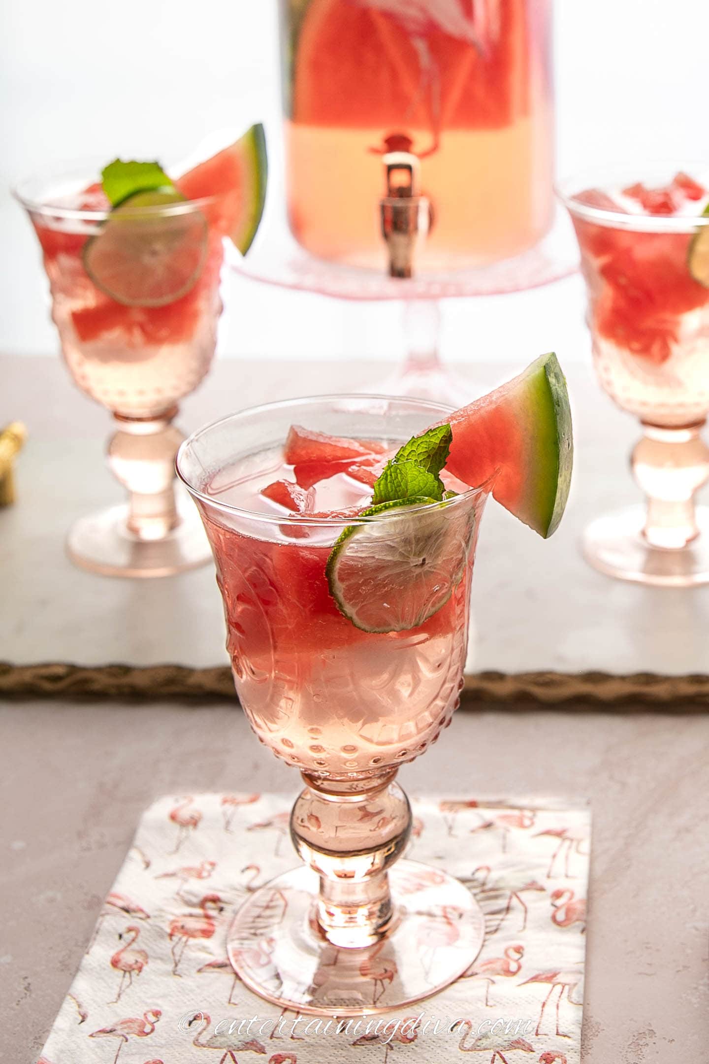a glass of watermelon sangria in front of a drink dispenser and two other glasses