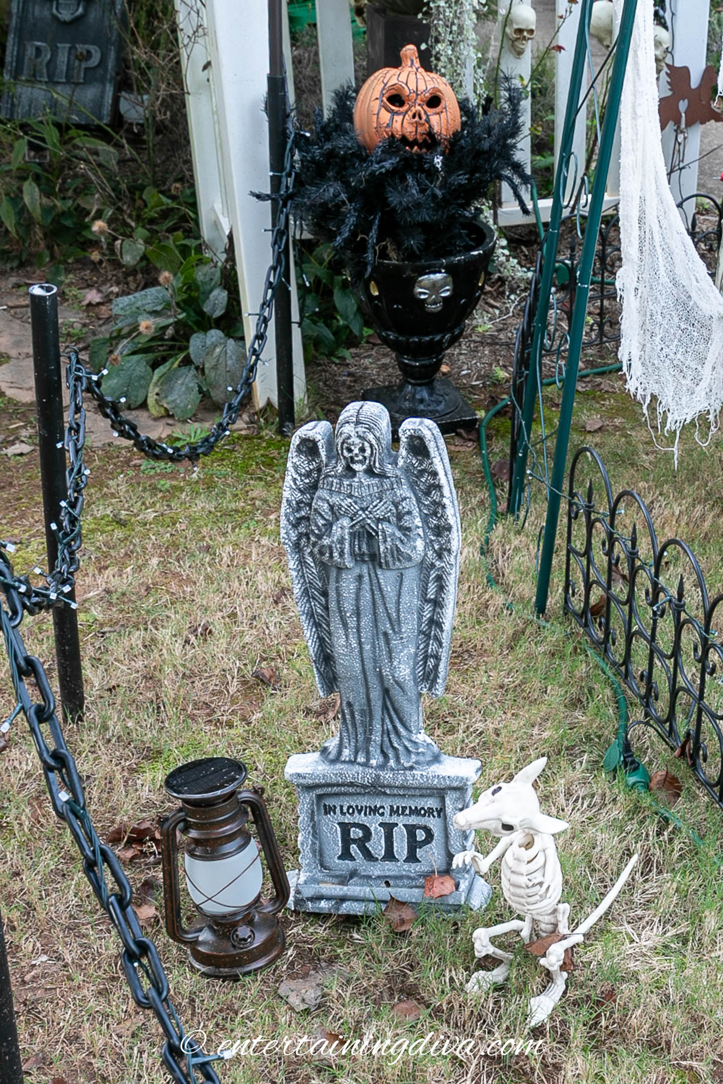 A Halloween yard display featuring an angel tombstone, skeleton dog, lantern, black urn with a pumpkin head, and black fence on grass.
