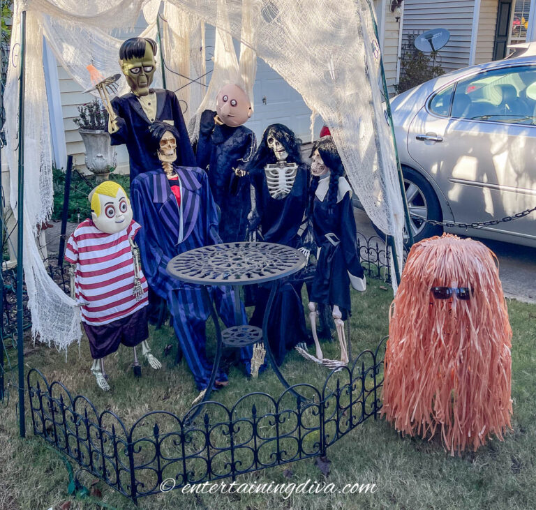 A group of Halloween decorations resembling cartoon and skeleton characters is arranged around a metal table and chairs in a yard.