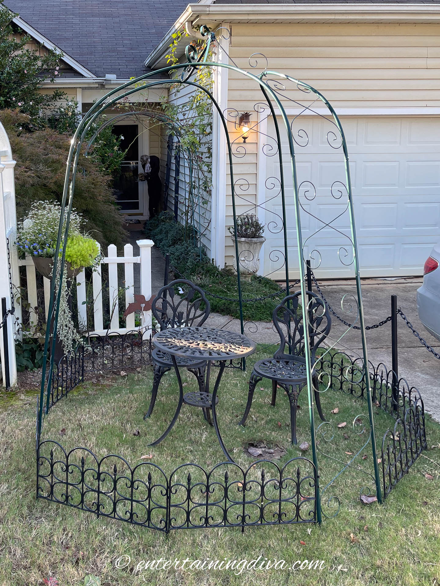 A small metal gazebo frame with two black metal chairs and a round table sits on a lawn in front of a house and white picket fence.