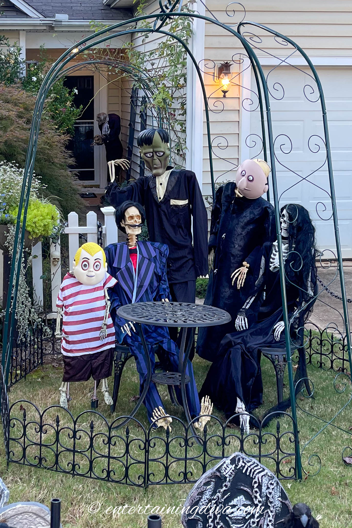 Five Halloween skeleton and monster figures are arranged around a small metal table in a fenced yard, set up as a spooky outdoor scene.
