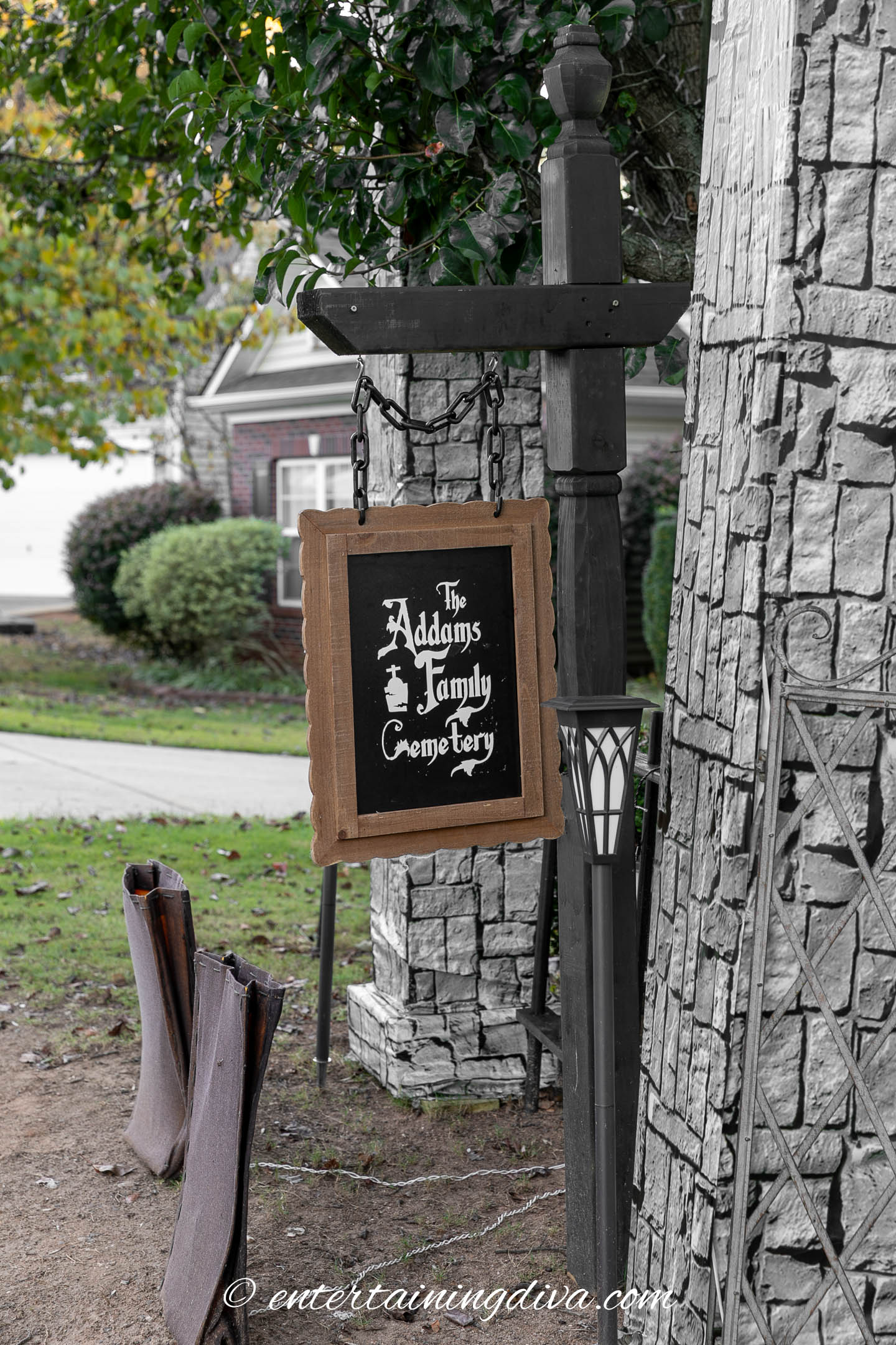 A sign reading "The Addams Family Cemetery" hangs from a black post in a yard decorated with artificial stone and Halloween props.