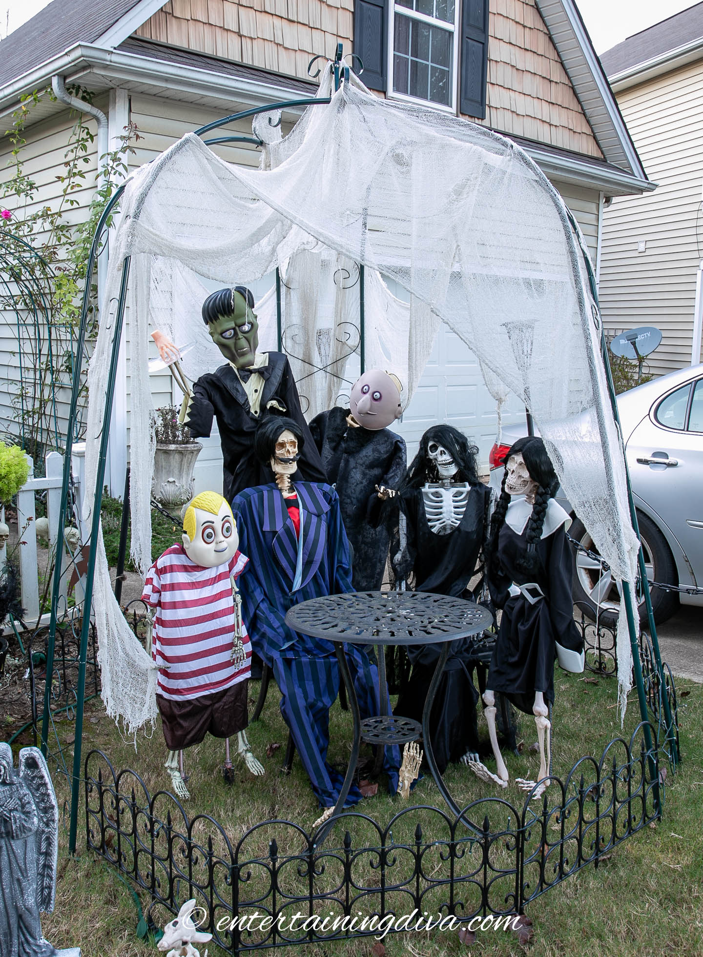 A Halloween yard display features life-sized skeleton and monster figures in costumes, arranged around a small black table under a white draped canopy.