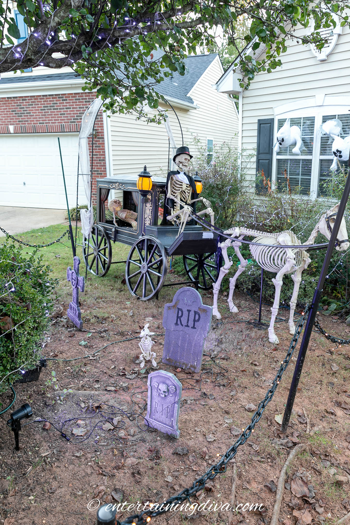 Halloween yard display featuring a skeleton in a horse-drawn hearse, tombstones labeled "RIP," and decorative skeleton horse in front of a house with ghost decorations in the window.