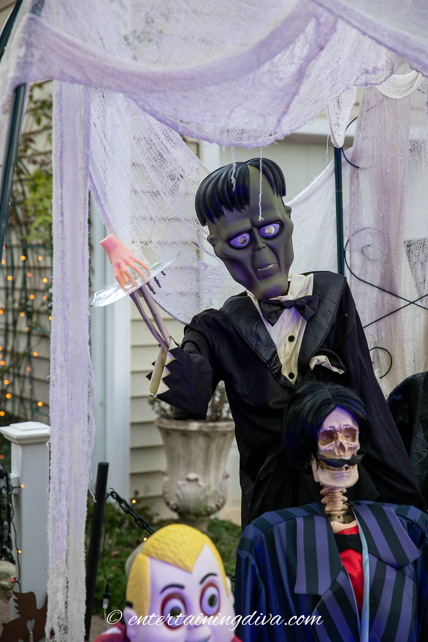 Three Halloween skeleton decorations dressed in costumes, including one in a tuxedo with purple eyes holding a tray, set in front of a house under spiderweb decorations.