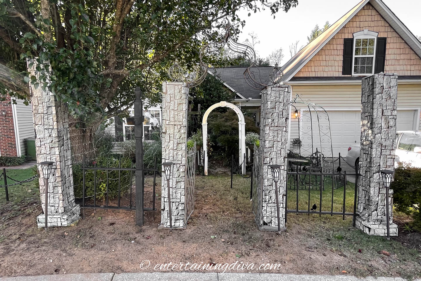 Stone columns and black metal fencing enclose a small front yard with a white garden arch, beside a house with shingle siding and a two-car garage.