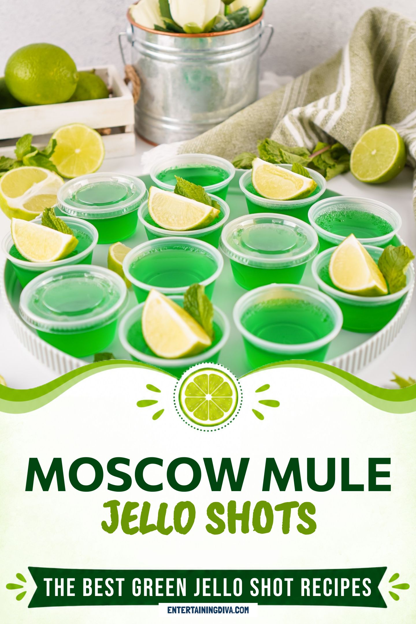 A plate of green Moscow Mule Jello shots in plastic Jello shot cups, garnished with a lime wedge and mint leaves. Limes, mint, and a towel are in the background.