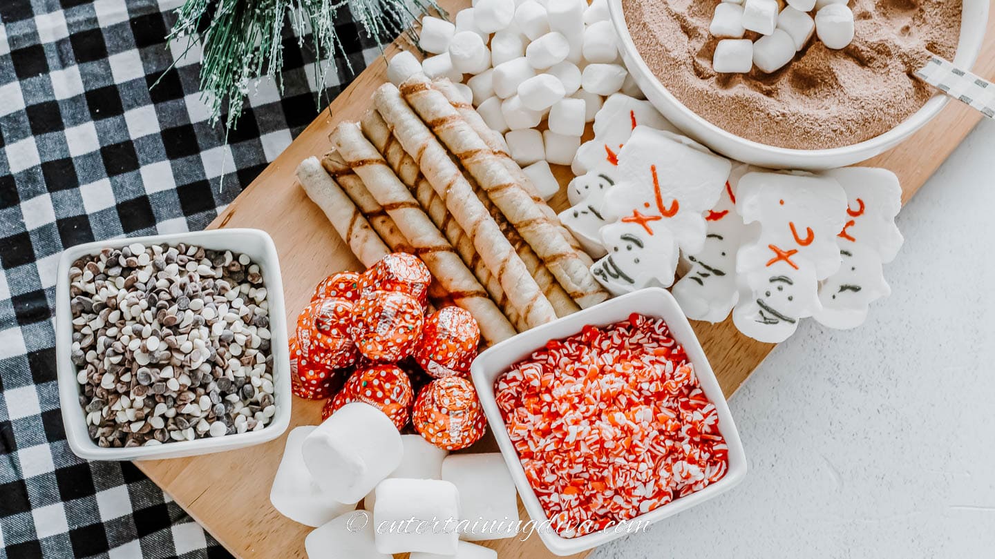 hot chocolate mix inn a bowl, marshmallows, snowman peeps, wafer cookies, crushed candy canes, and chocolate chips on a cutting board