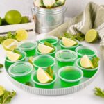 A plate of green Moscow Mule Jello shots in plastic Jello shot cups, garnished with a lime wedge and mint leaves. Limes, mint, and a towel are in the background.