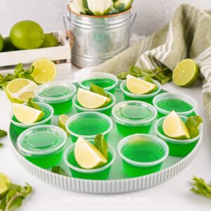 A plate of green Moscow Mule Jello shots in plastic Jello shot cups, garnished with a lime wedge and mint leaves. Limes, mint, and a towel are in the background.