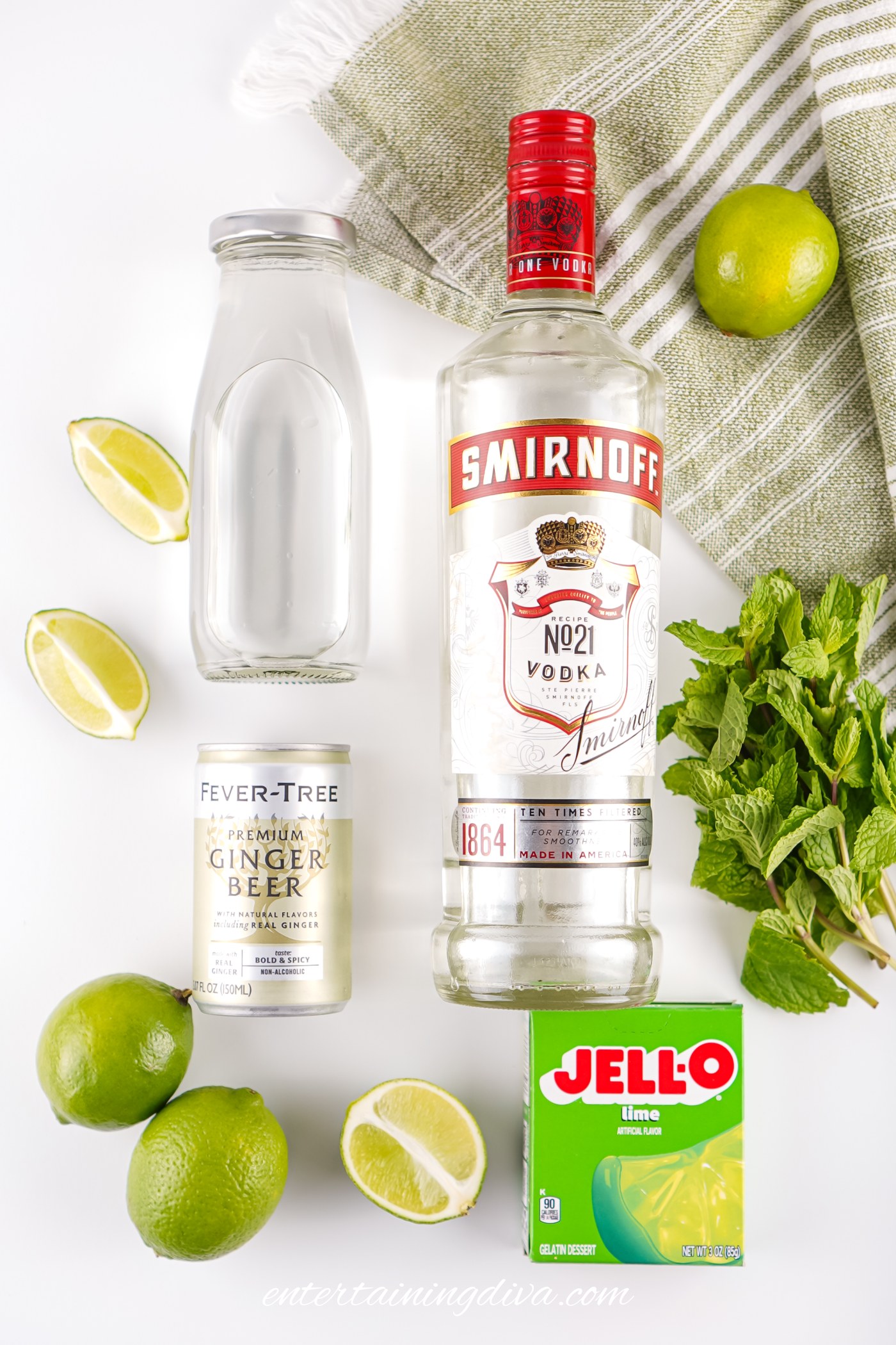 Top-down view of a Smirnoff vodka bottle, a bottle of water, a can of ginger beer, lime Jello, fresh lime, lime wedges, mint, and a green-striped towel.
