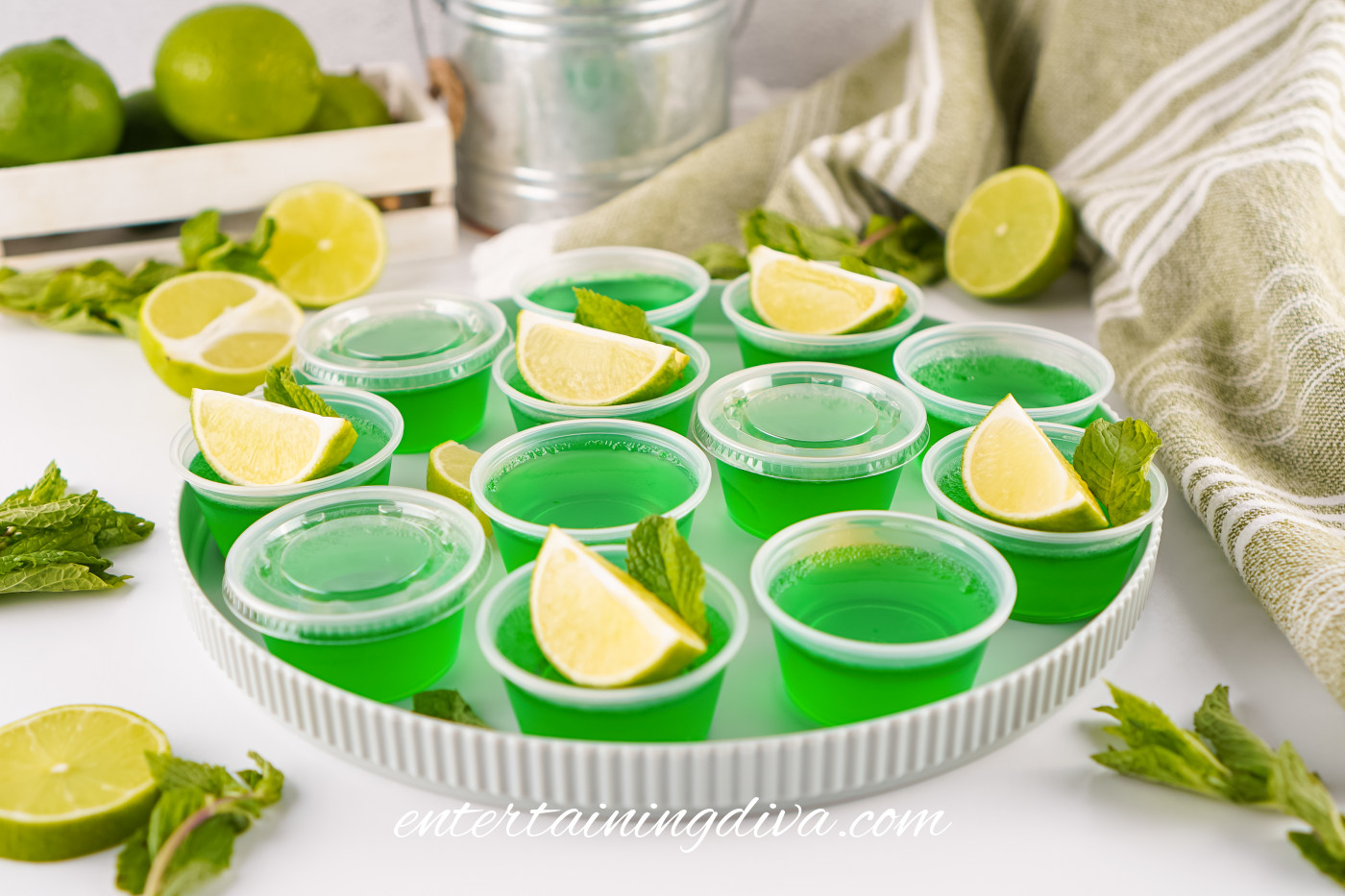 A round tray holding green Jello shots in plastic cups, each garnished with a lime wedge and mint leaves; whole and halved limes and a green towel are nearby.
