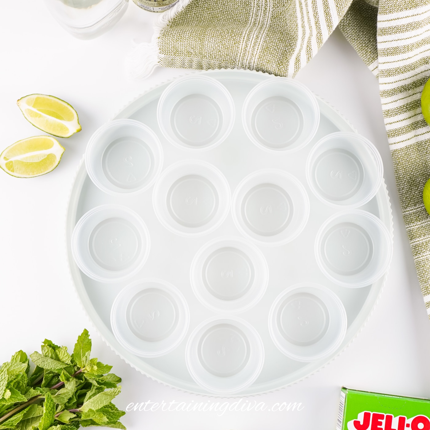 A round white tray holds twelve empty Jello shot cups, surrounded by lime wedges, mint leaves, a green-striped towel, and a box of Jello.