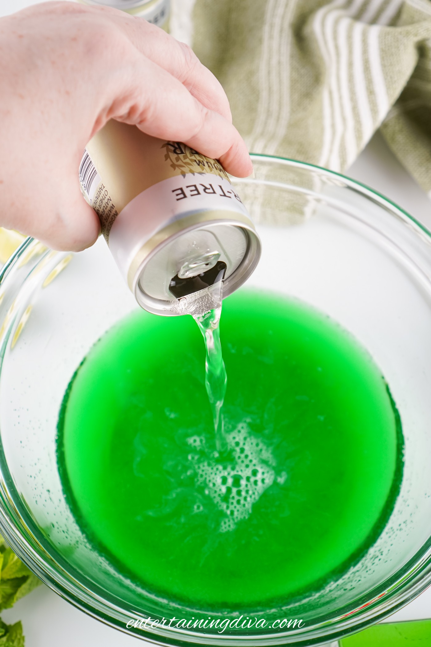 A hand pours ginger beer from a can into a glass bowl filled with green Jello mix.