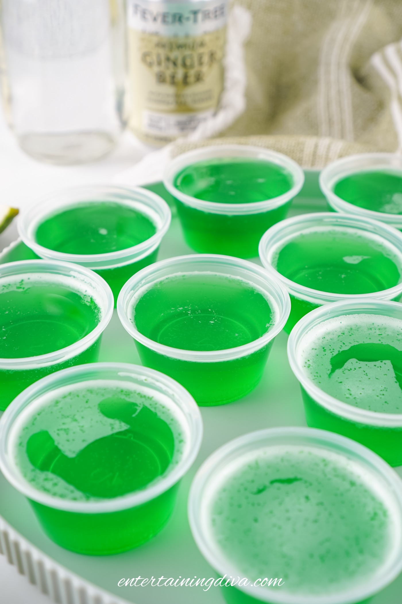 Several plastic Jello shot cups filled with bright green Moscow Mule Jello shots are arranged on a white surface, with bottles and a towel in the background.