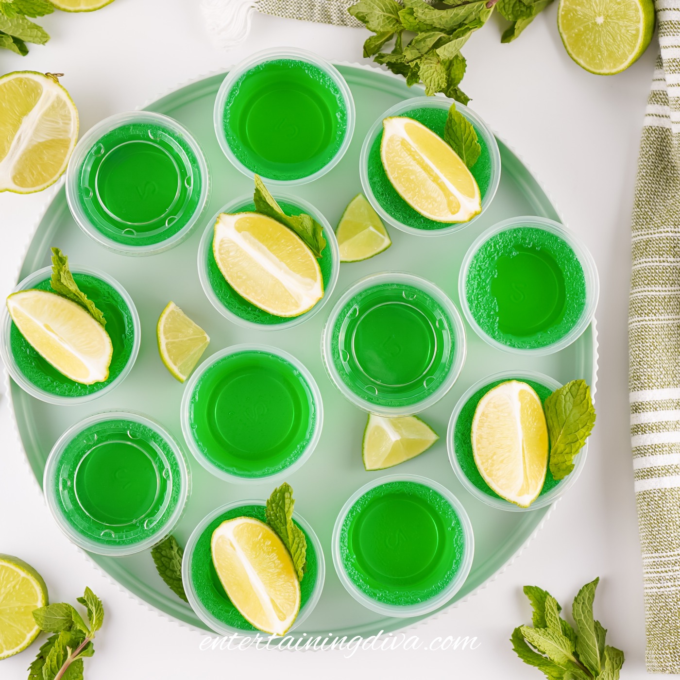 Overhead view of a round tray with green Moscow Mule Jello shots in plastic cups, garnished with a lime wedge and fresh mint; extra limes and mint scattered around.