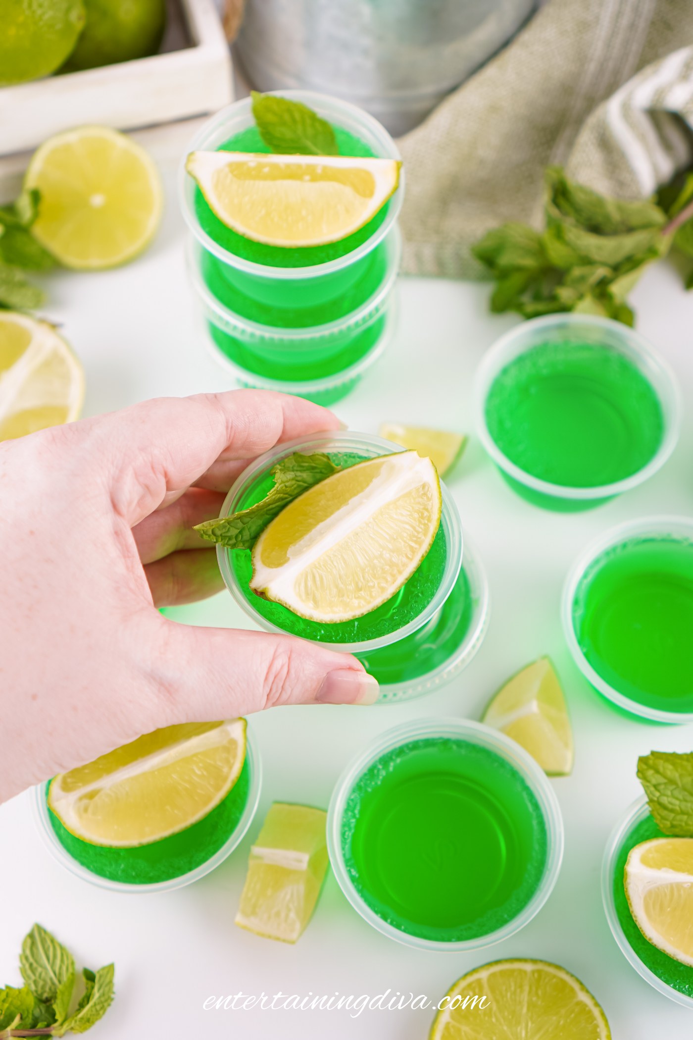 A hand holds a small Jello shot cup filled with a Moscow Mule Jello shot and garnished with a lime wedge and mint; several more shots are on a white surface surrounded by lime slices and mint leaves.