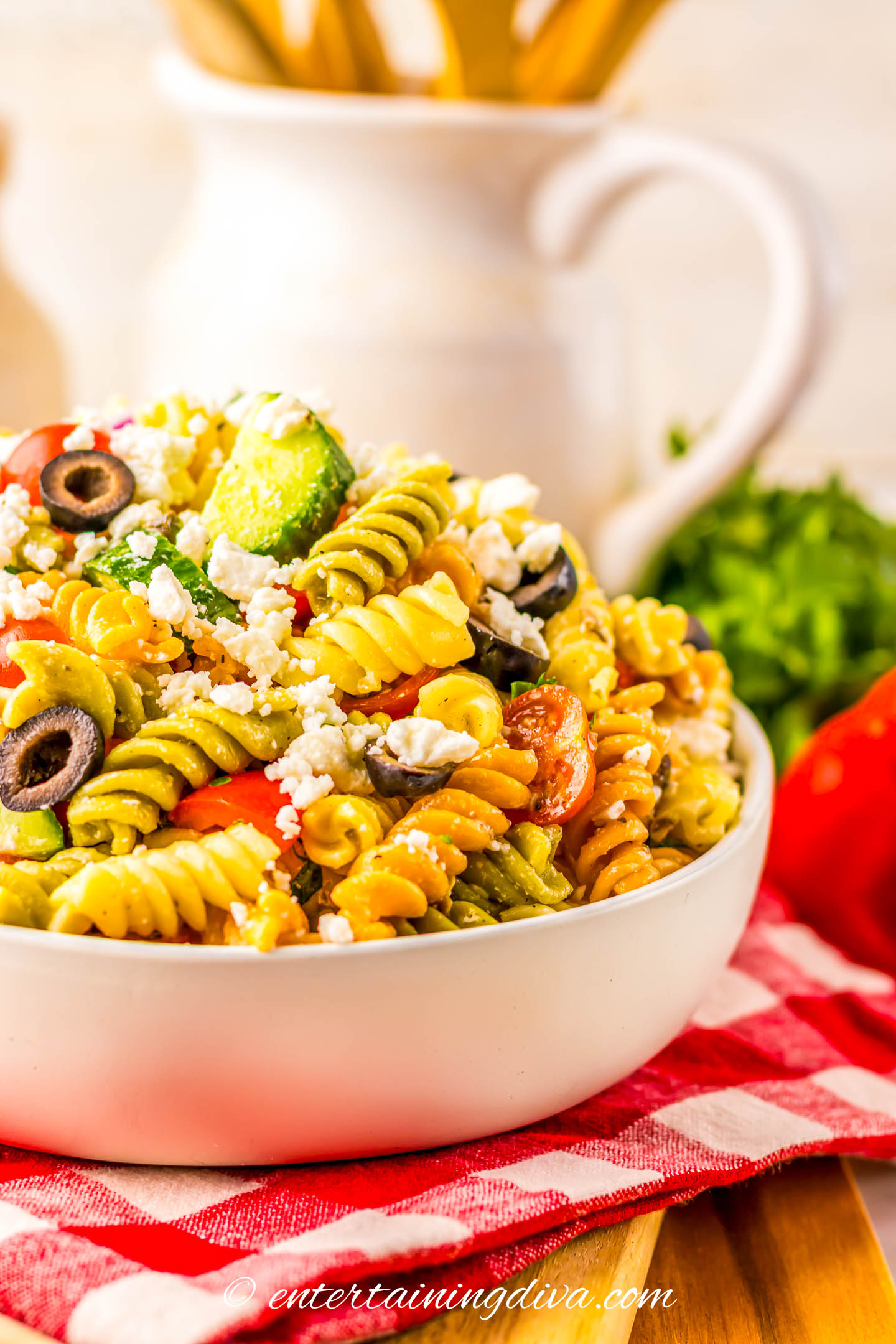 A bowl of tri-color rotini pasta salad with cherry tomatoes, black olives, cucumbers, and crumbled feta cheese on a red checkered cloth.