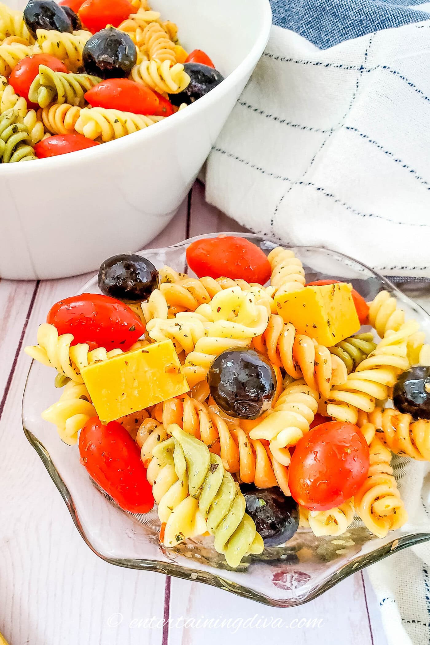 A bowl and a small dish of tri-color rotini pasta salad with cherry tomatoes, black olives, cheddar cheese cubes, and Italian seasoning.