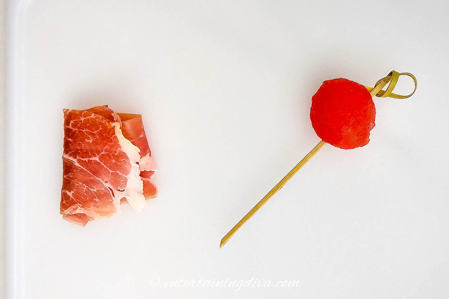 A folded slice of prosciutto beside a watermelon ball on a skewer