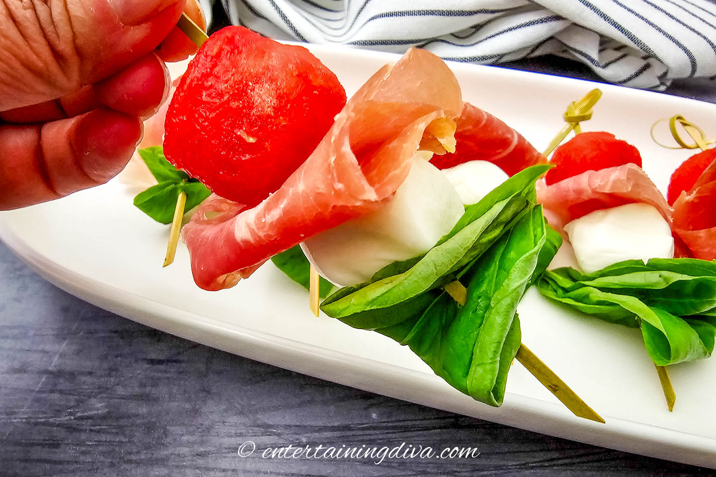 A close up of a watermelon prosciutto skewer being picked up from a tray