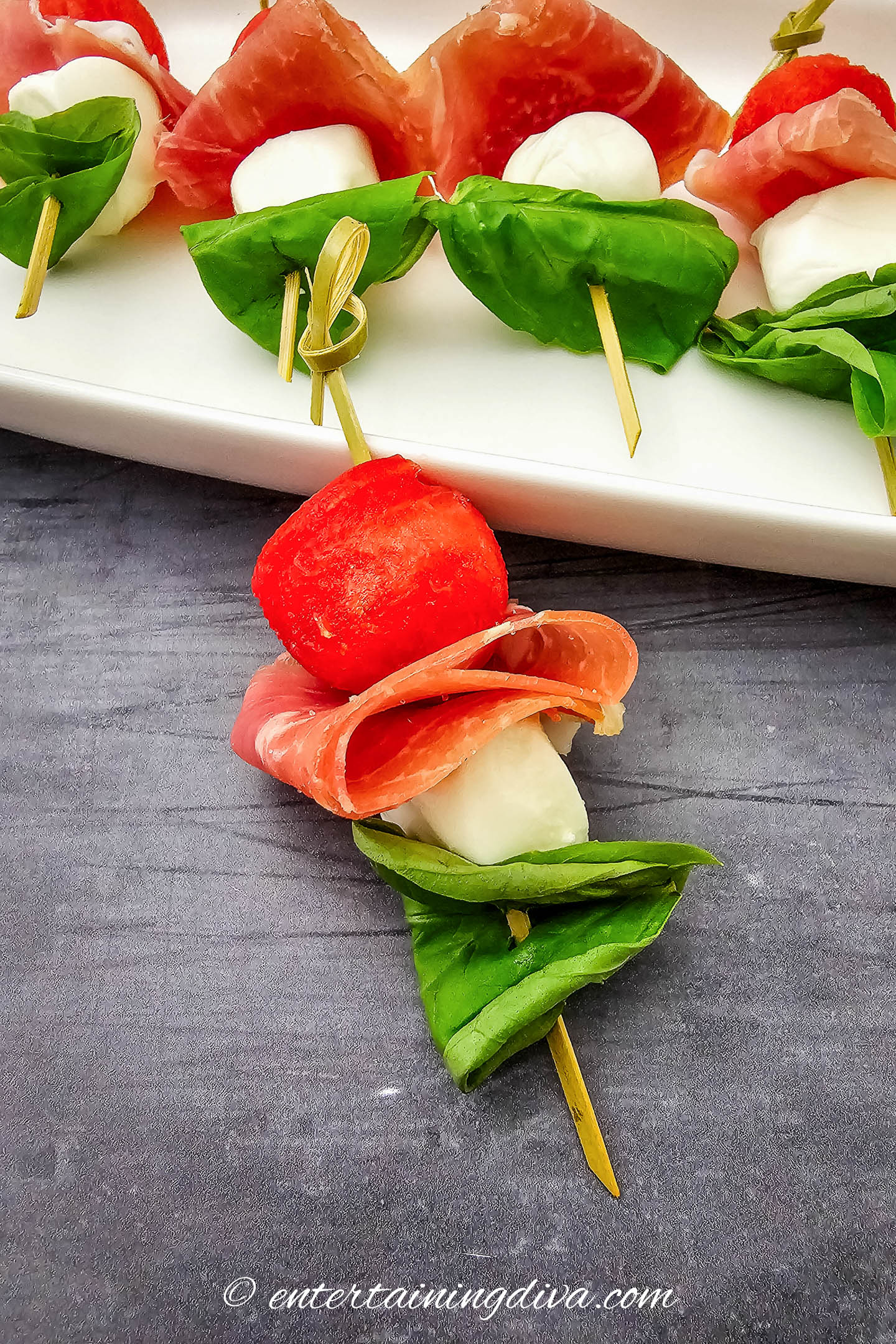 A watermelon caprese skewer on a countertop beside a tray with more skewers