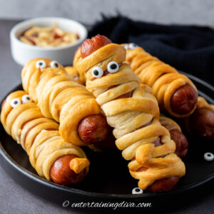 A plate of crescent roll-wrapped hot dogs decorated with candy eyes to resemble mummies, with a bowl of dipping sauce in the background.