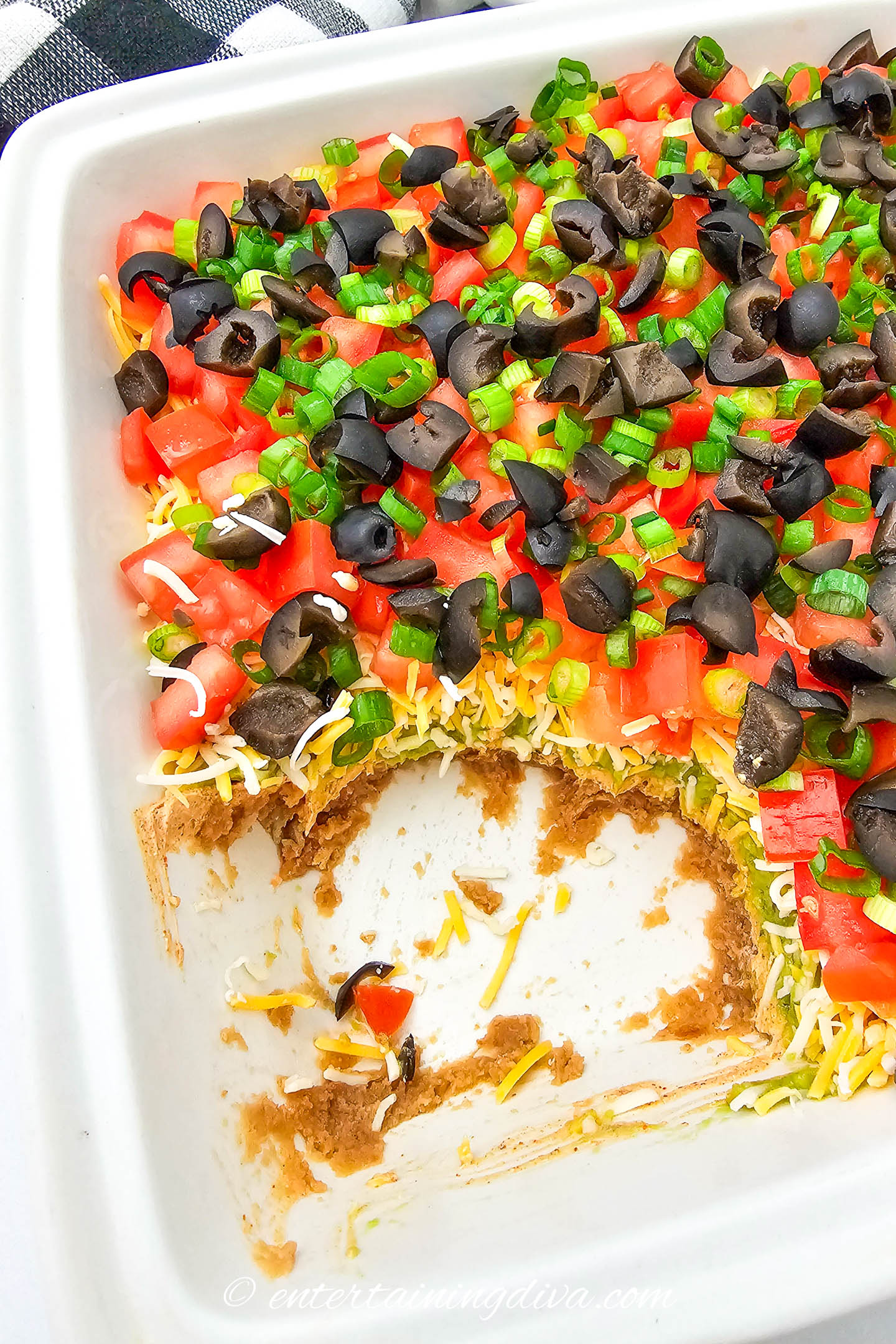 Rectangular dish of layered dip with a portion missing, showing layers of refried beans, cheese, diced tomatoes, black olives, and chopped green onions.
