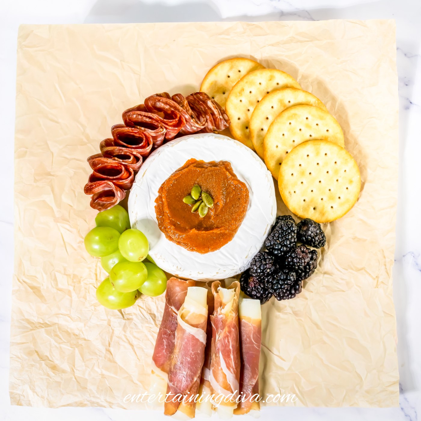 A charcuterie board with brie, pumpkin butter, salami, crackers, prosciutto wrapped mozzarella, green grapes and blackberries