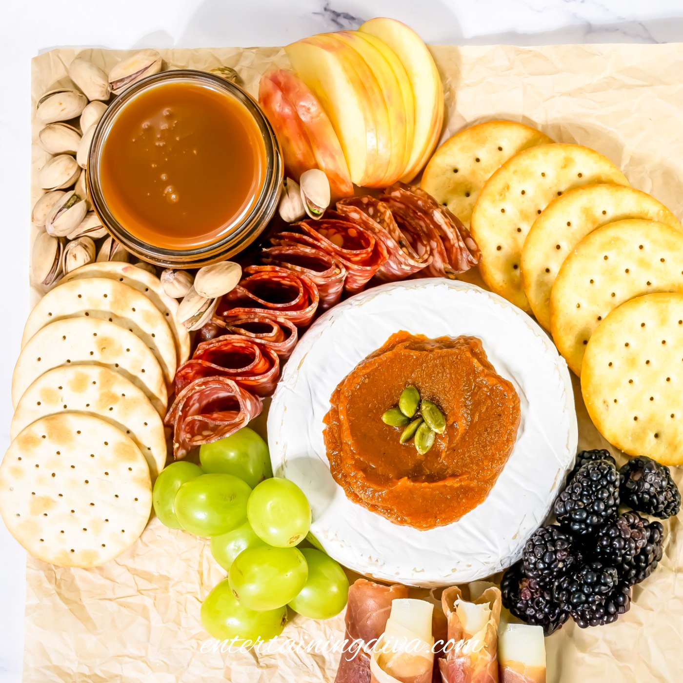 A corner of a charcuterie board with brie, pumpkin butter, salami, crackers, prosciutto wrapped mozzarella, green grapes, blackberries, caramel sauce, apple slices and pistachios