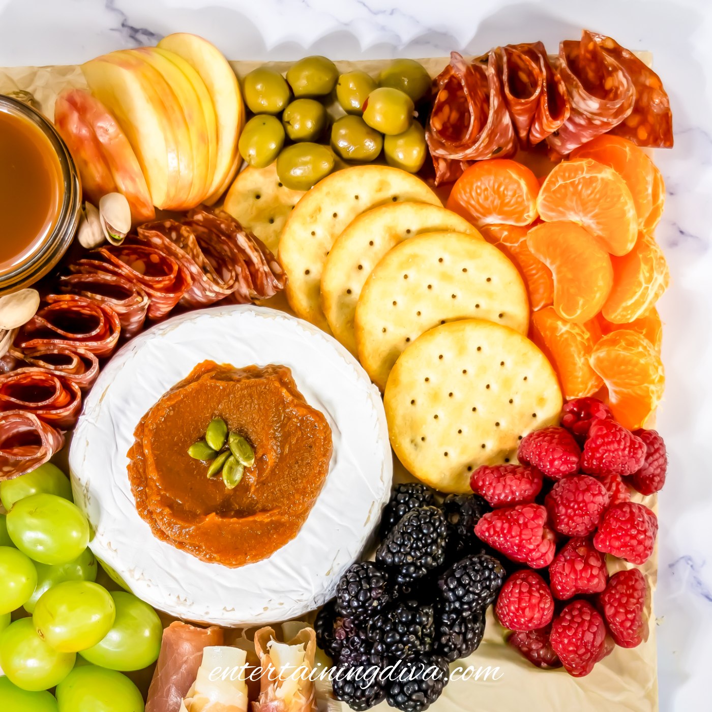 The top corner of a fall charcuterie board with brie, pumpkin butter, salami, sliced apples, olives, crackers, mandarin orange slices, raspberries and blackberries
