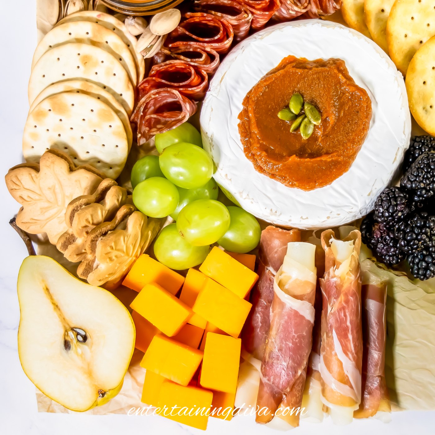 The bottom left corner of a Charctuerie board with half of a pear, maple leaf cookies, cubed cheddar cheese, green grapes, salami, brie, and crackers