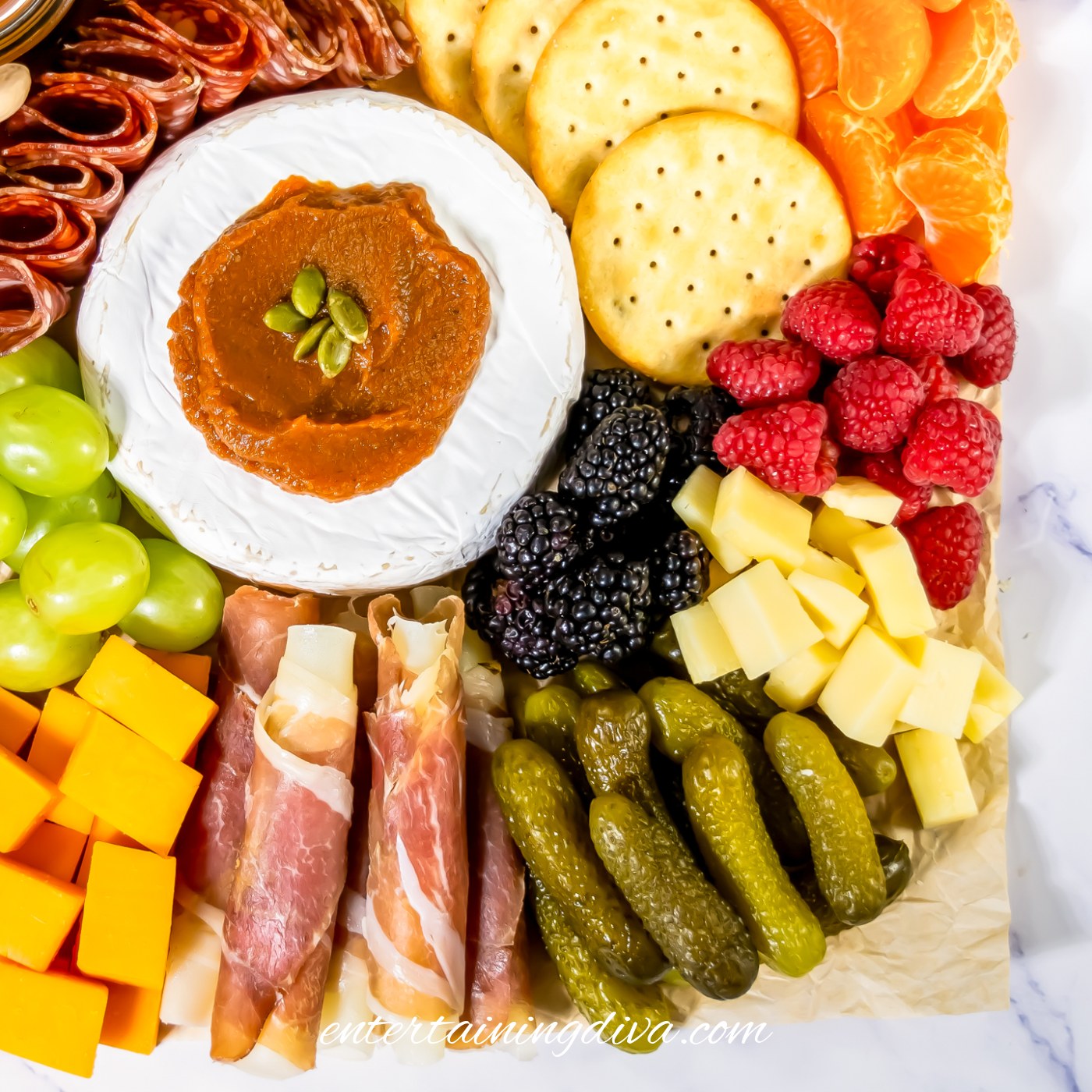 The bottom right corner of a Charcuterie board with mini pickles, cubed white cheese, raspberries, crackers, meat wrapped mozzarella and brie