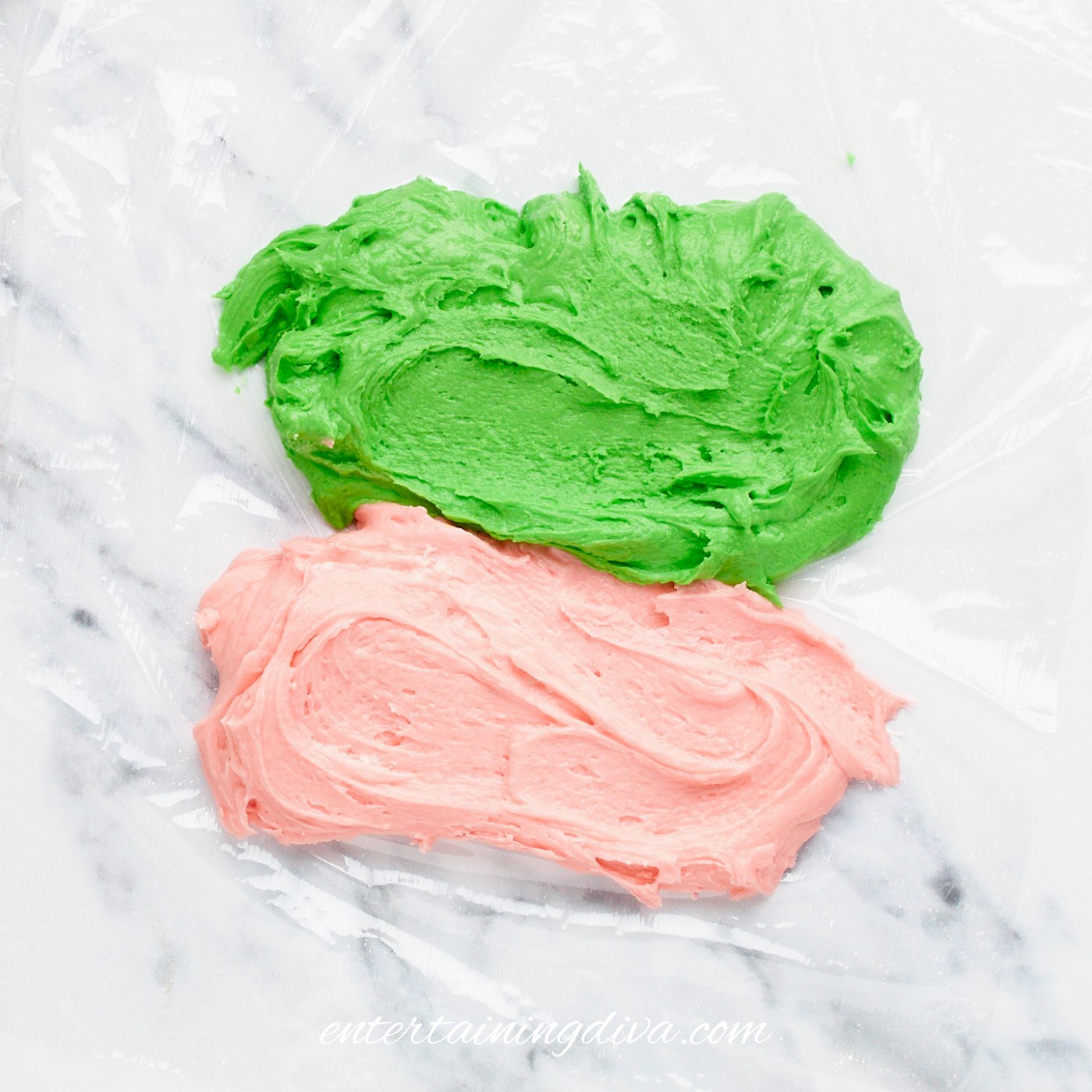 A row of green buttercream frosting beside a row of pink buttercream frosting on plastic wrap.