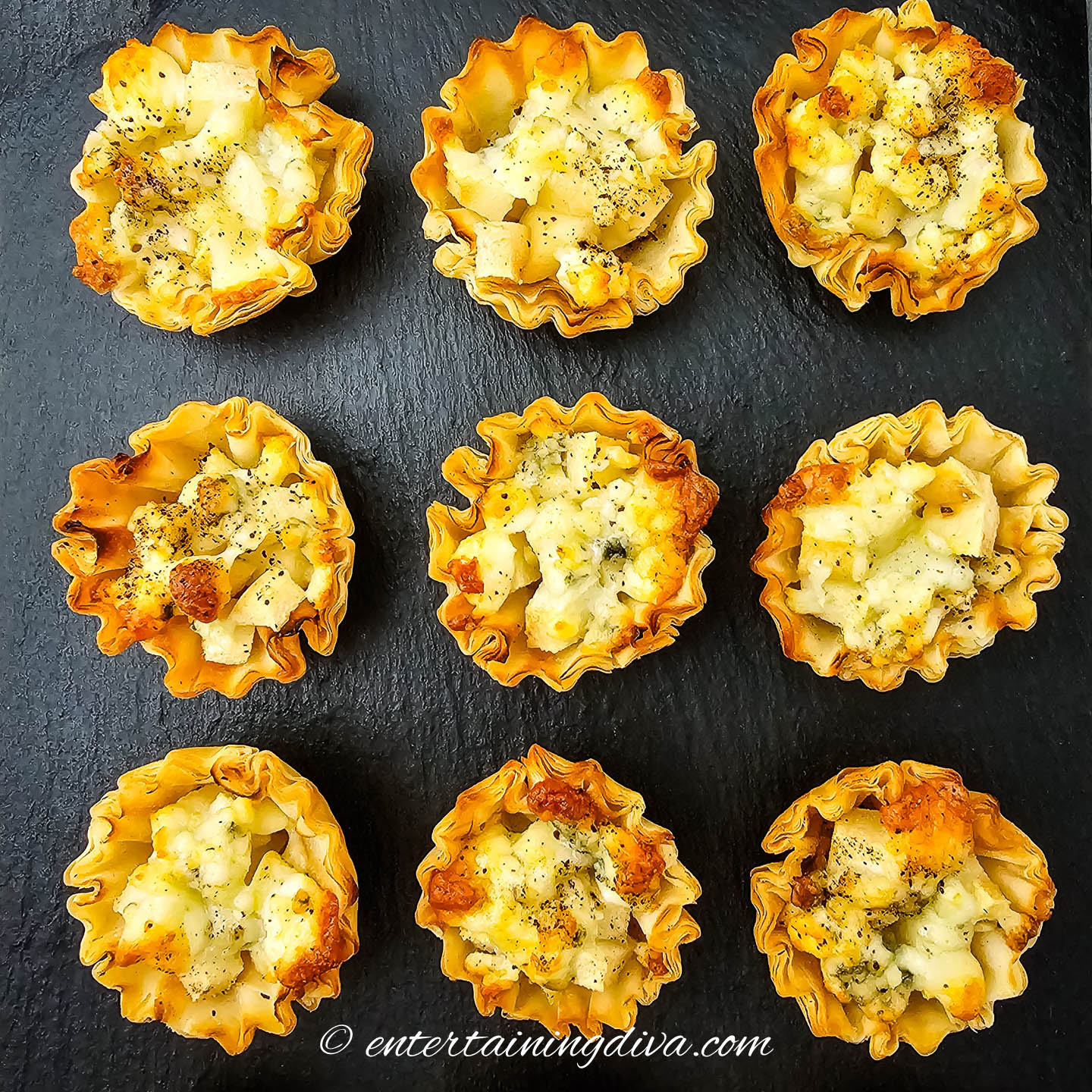 Nine phyllo pastry cups filled with a diced potato and cheese mixture, arranged in a grid on a black slate surface.