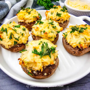 Five baked stuffed mushrooms topped with melted cheese and fresh parsley are arranged on a white plate, with a small bowl of grated cheese in the background.