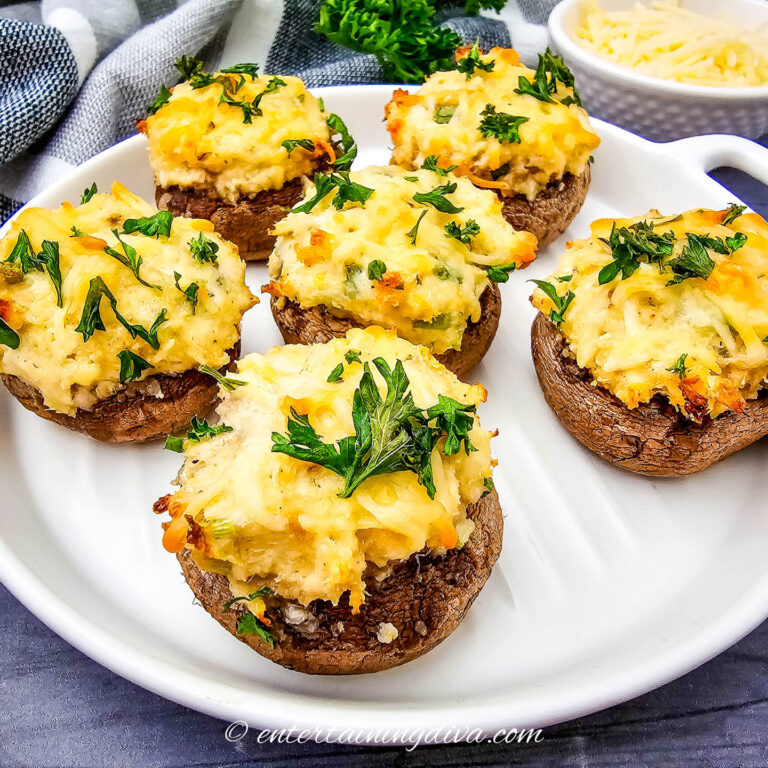 Five baked stuffed mushrooms topped with melted cheese and fresh parsley are arranged on a white plate, with a small bowl of grated cheese in the background.